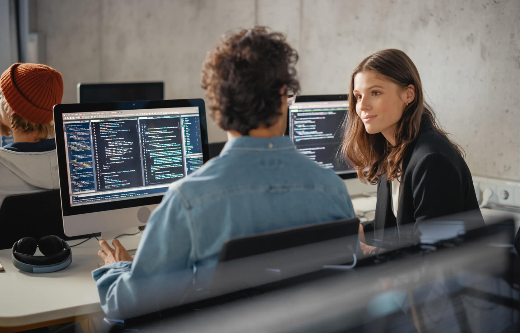 Man and a woman talking and sitting in front of computer monitors displaying code on the screens