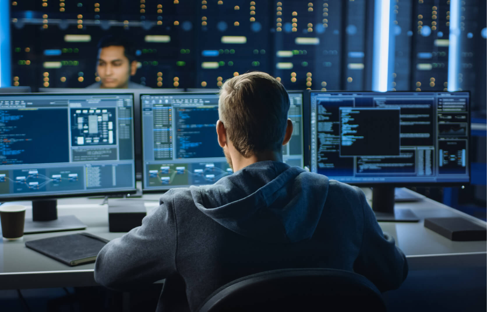 A male cybersecurity professional sitting in front of a computer with three monitors as he conducts dark web monitoring