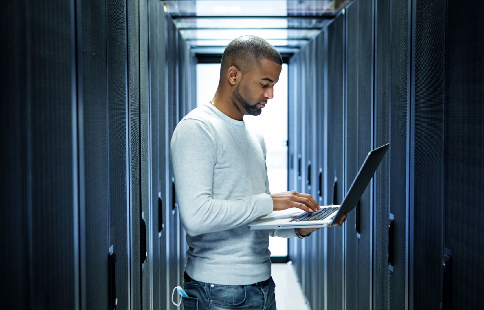 A male IT professional standing in the hallway of a server room working on a laptop