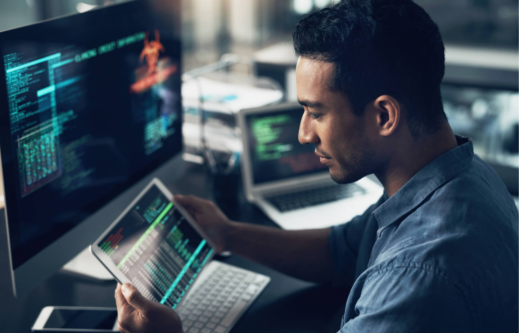 A male virtual CISO sits at desk with a laptop and large monitor, while looking at data on a small handheld tablet