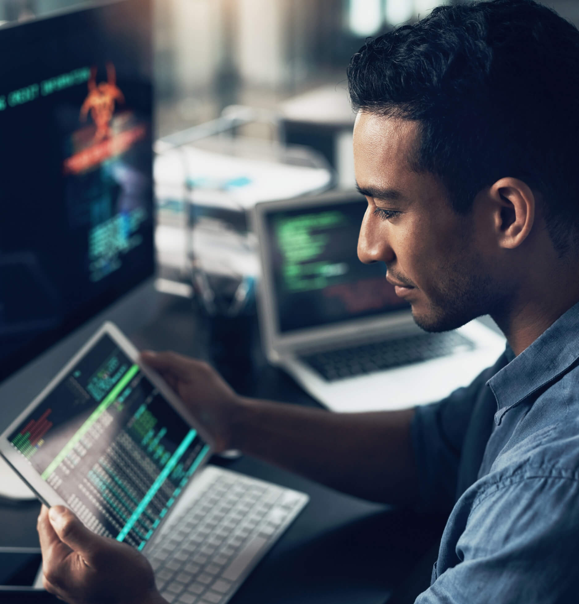 A male virtual CISO sits at desk with a laptop and large monitor, while looking at data on a small handheld tablet